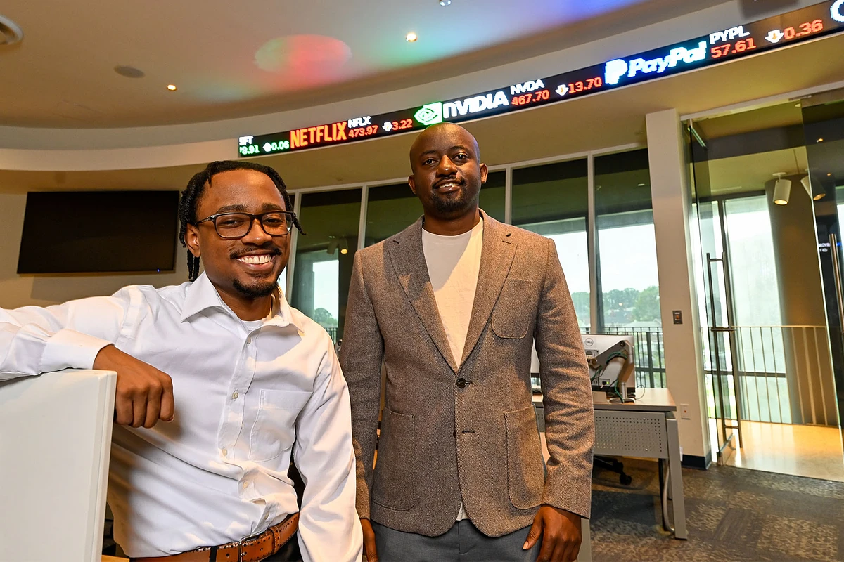 Two men stand smiling in an office environment with a digital stock ticker in the background.