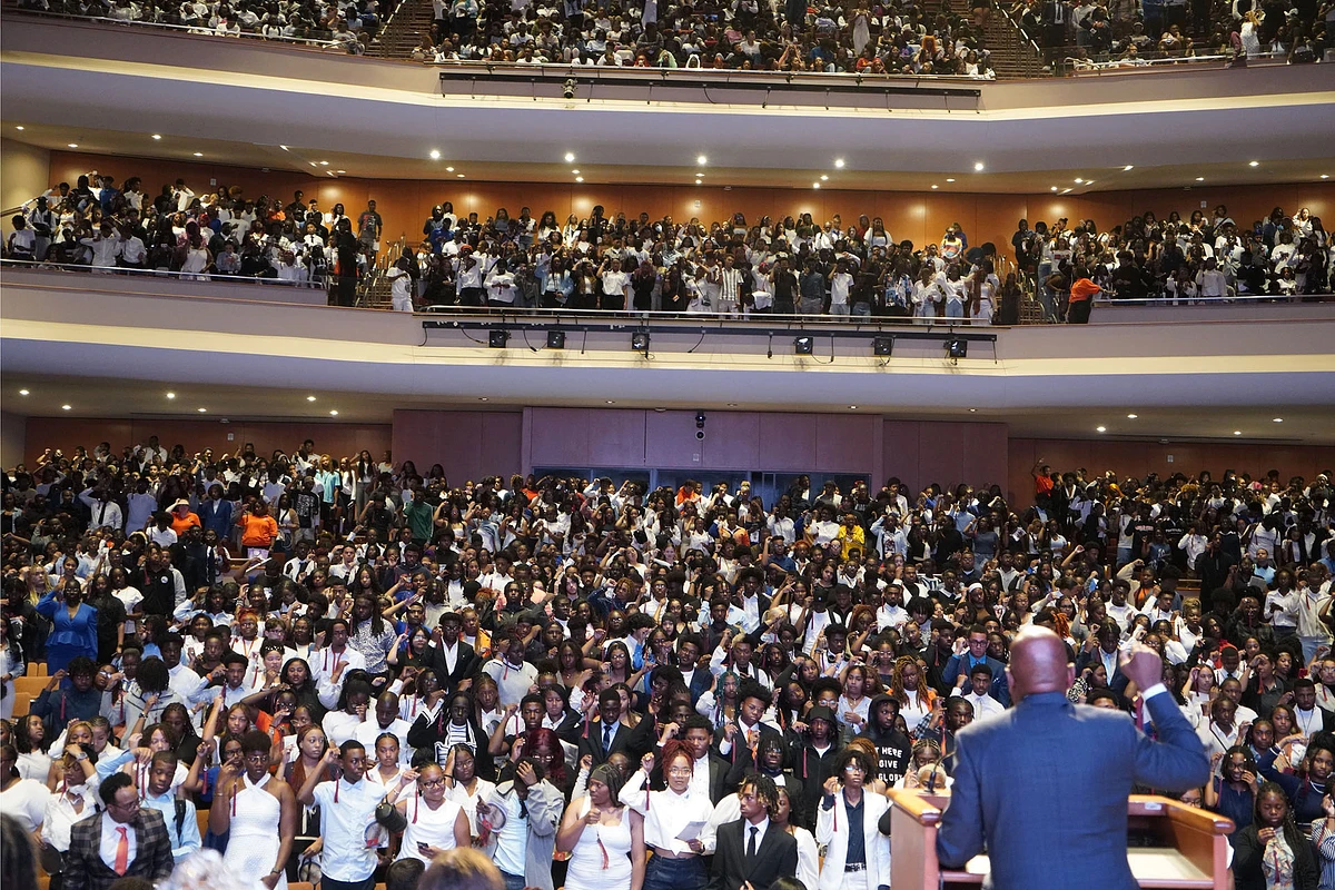 A large crowd of people, many wearing formal attire, gather in an auditorium as a speaker addresses them from a podium.